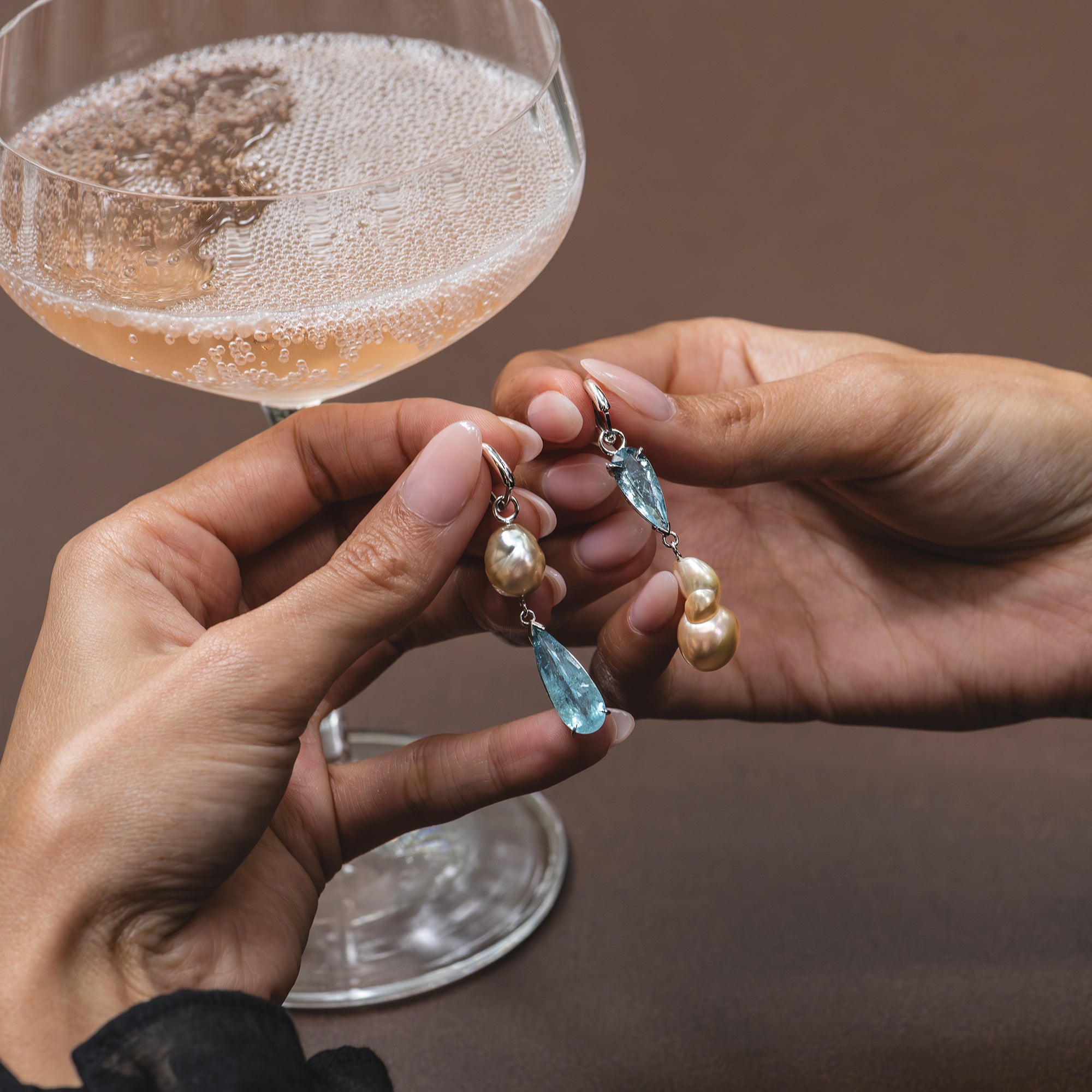 Aquamarine and golden pearl earrings held in a woman’s hands, sparkling rosé in the background.