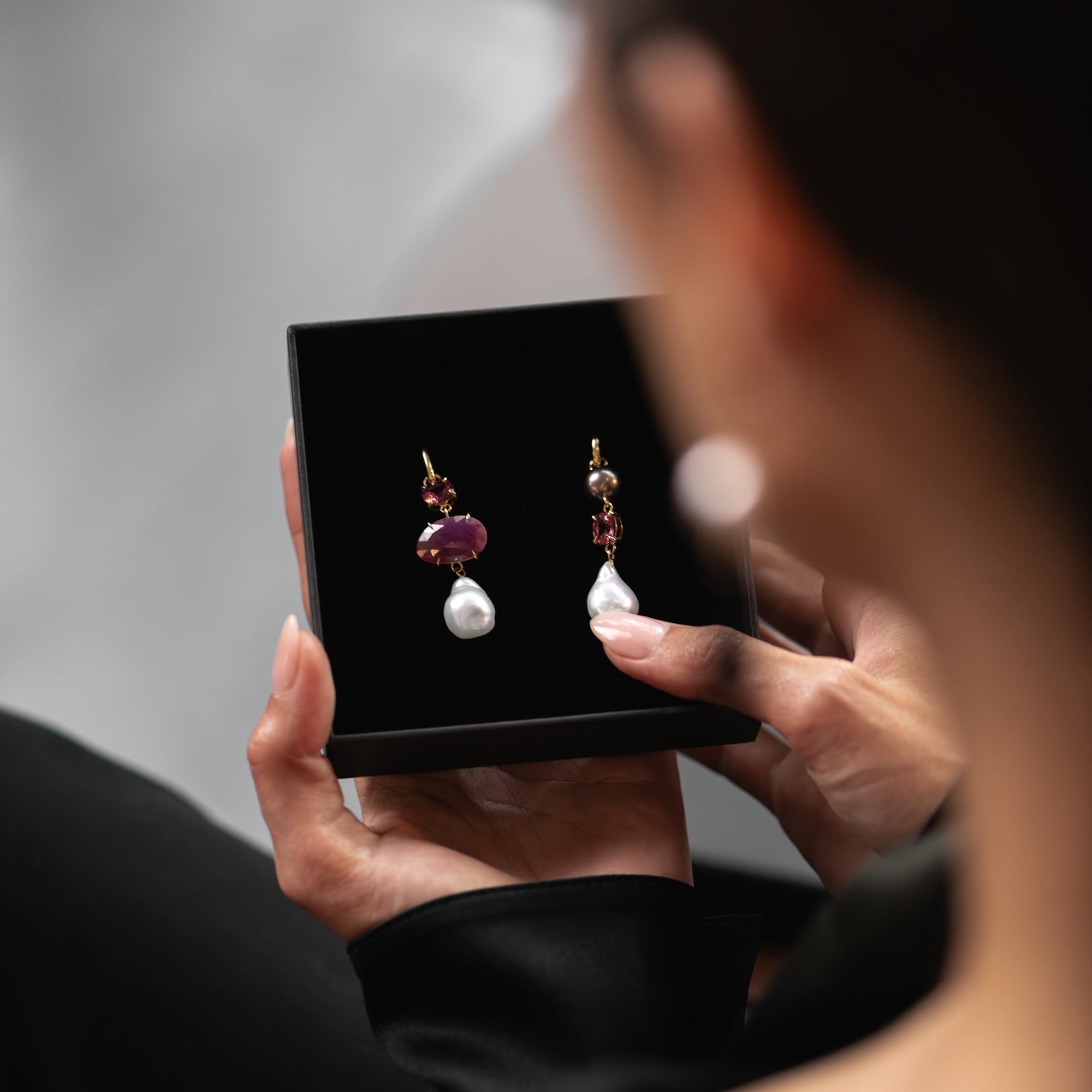A rose cut ruby, pink garnet, a purple Tahitian pearl, and white South Sea pearl earrings in black box being opened by a woman.
