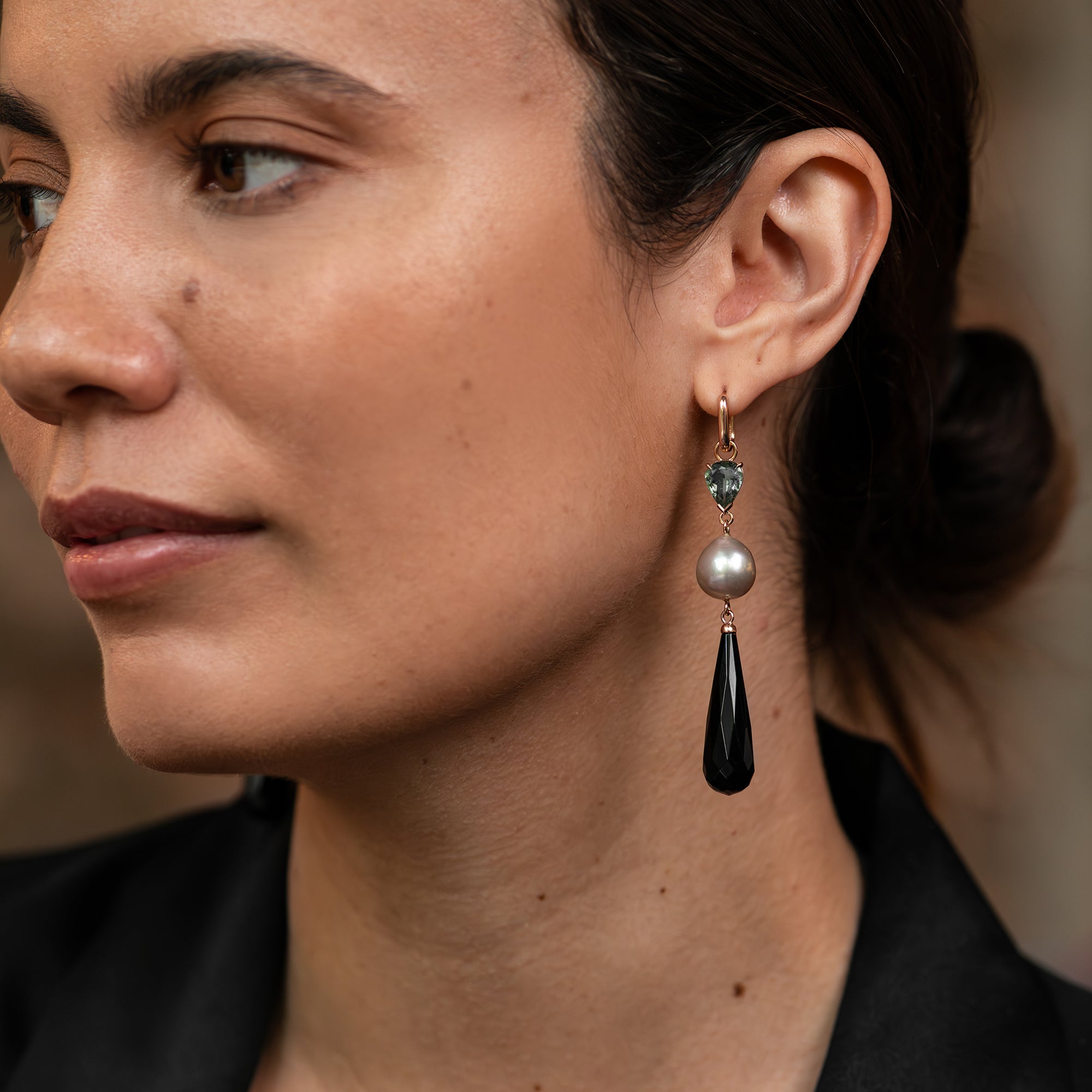 Rose gold huggie earring with pear cut green tourmaline, pink-hued South Sea pearl, and an onyx drop worn on a woman's ear.