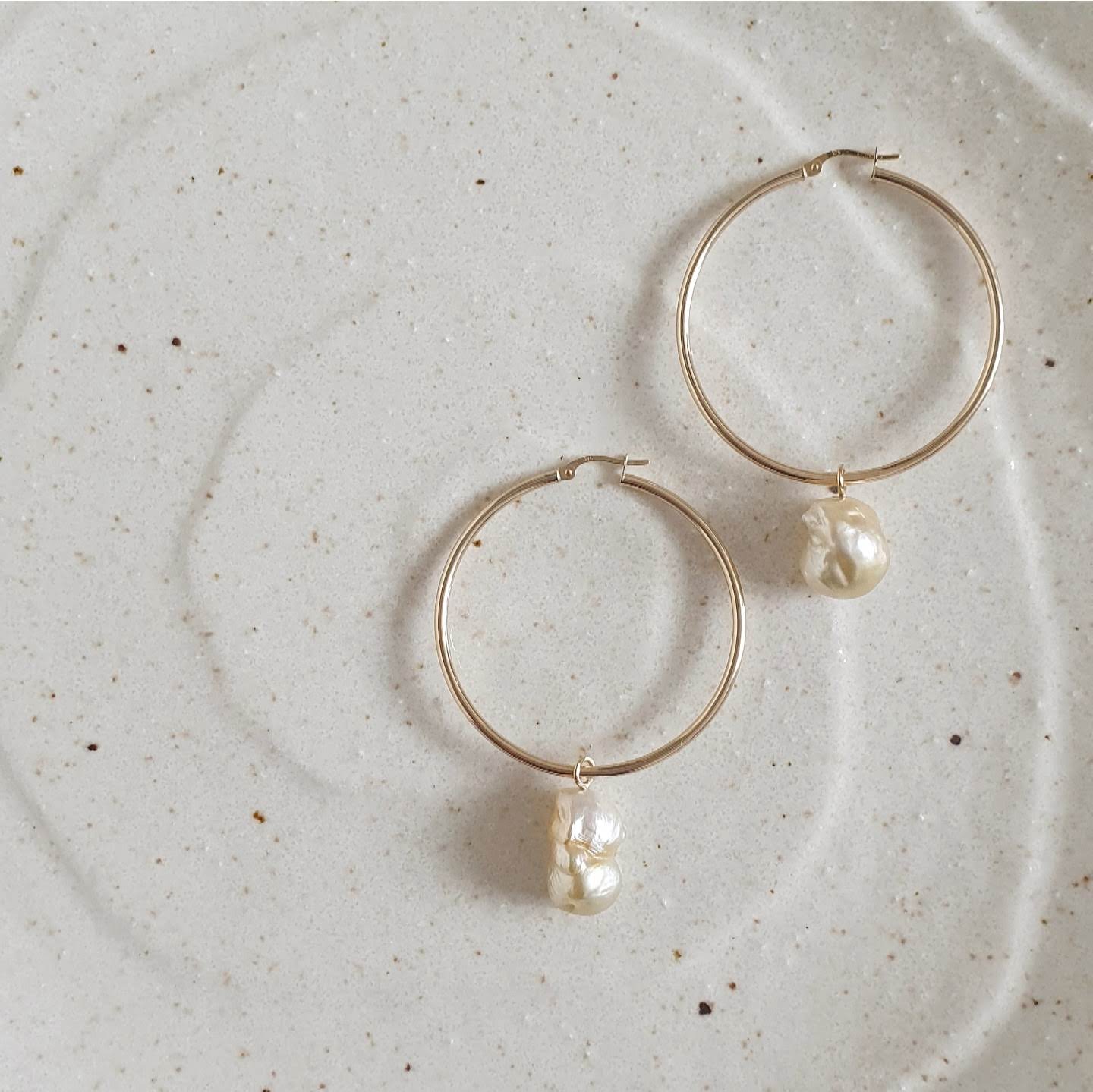 Hand selected unique baroque golden South Sea pearls, in a hand crafted yellow gold detachable setting, on large yellow gold high polish hoop earrings, displayed on a cream coloured textured ceramic platter.