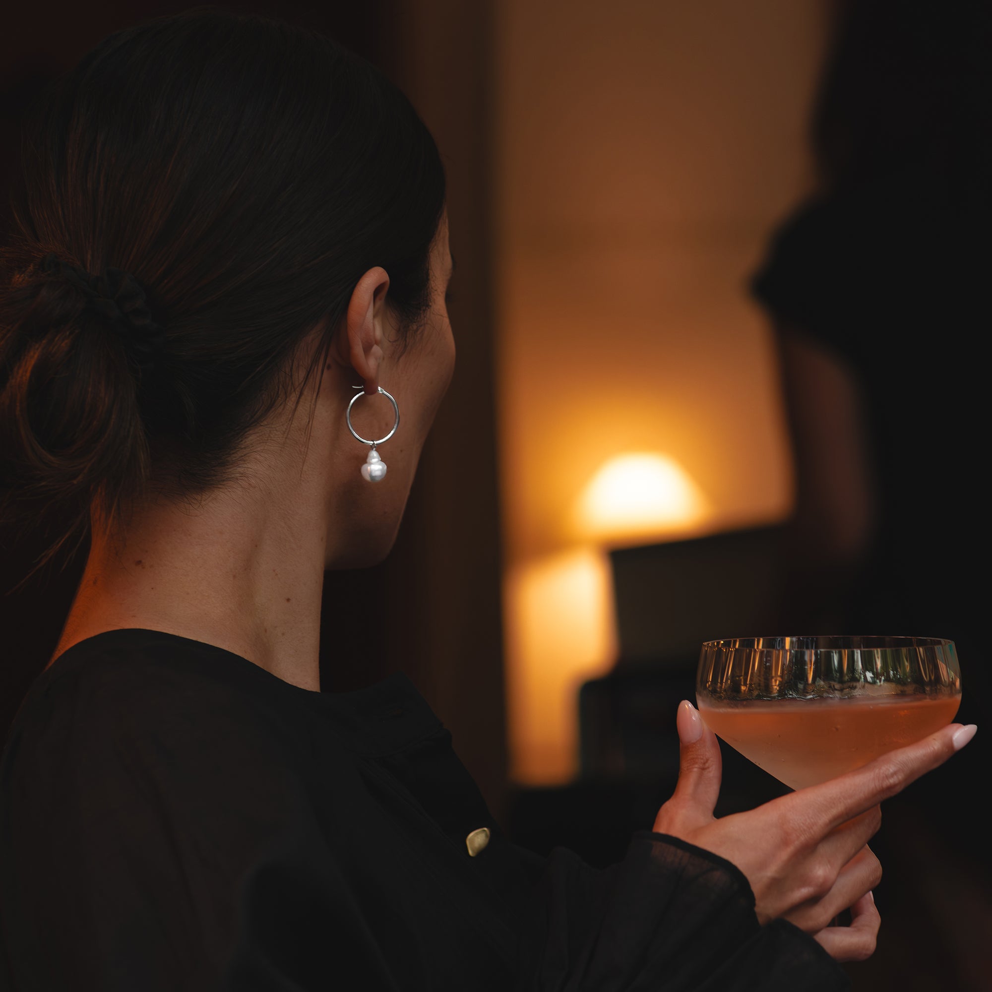Small white gold hoop earrings with detachable baroque white South Sea pearls, worn by a woman holding a coupe glass of sparkling rosé, facing away from the camera.