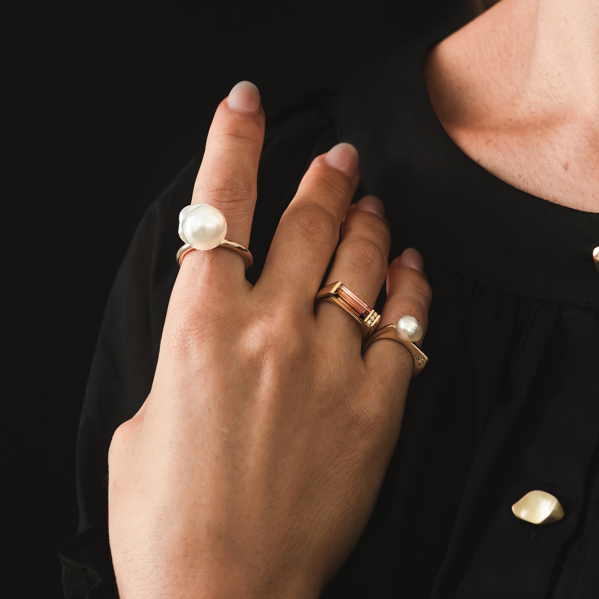 A yellow gold ring with high polish finish featuring a hand selected unique baroque white South Sea pearl and a trio of Australian champagne diamonds in earthy coloured tones is being worn on a woman's pinkie finger alongside a custom bespoke ring and an Ayla pearl ring..