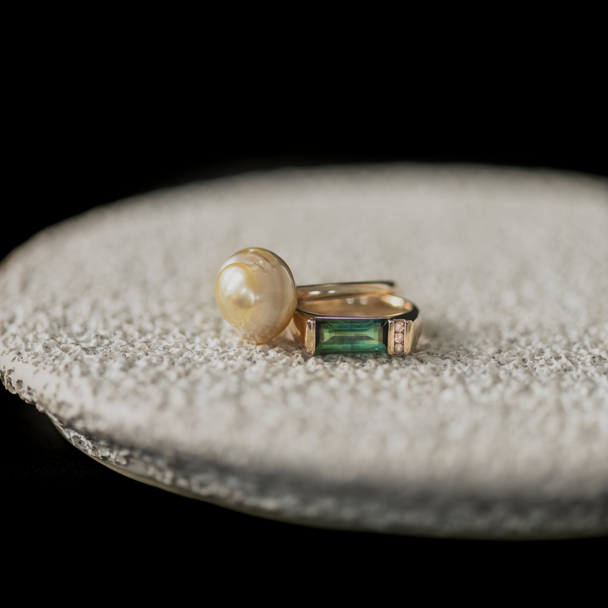 Yellow gold Ayla Pearl Ring with golden baroque South Sea pearl & custom bespoke ring with bi-colour green tourmaline & chocolate Australian champagne diamonds rest on a white textured ceramic dish.