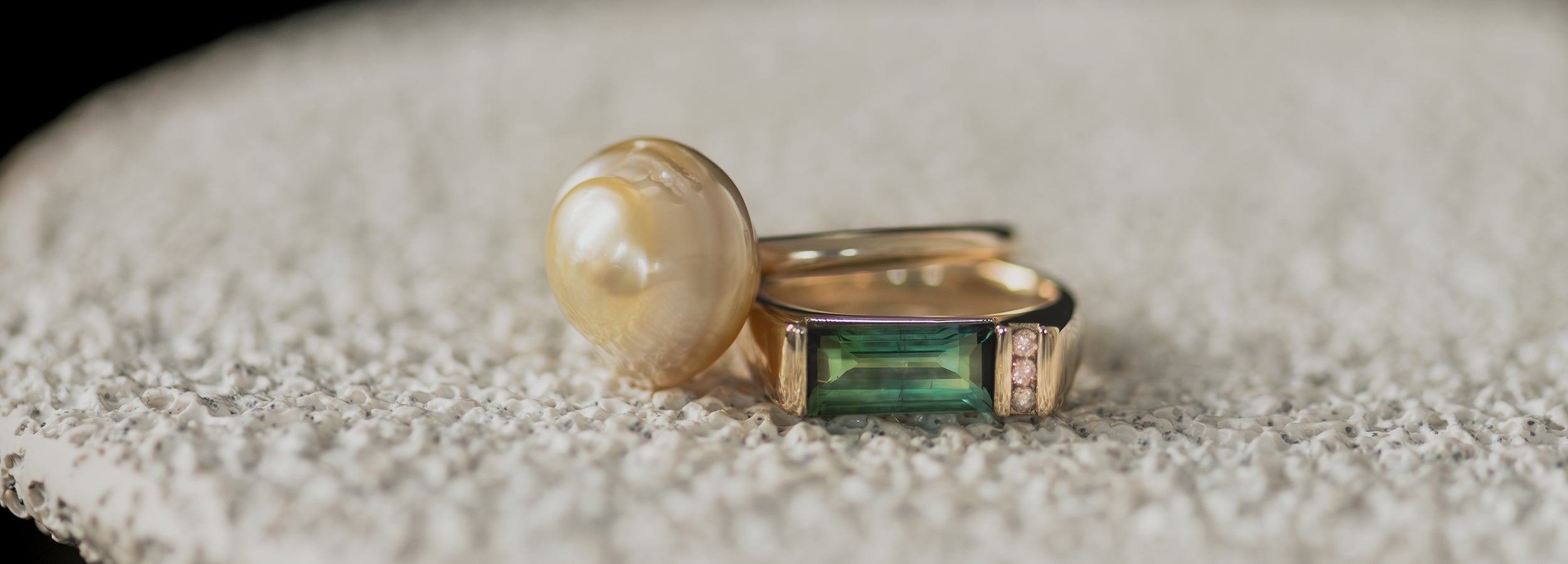 Yellow gold Ayla Pearl Ring with golden baroque South Sea pearl & custom bespoke ring with bi-colour green tourmaline & chocolate Australian champagne diamonds rest on a white textured ceramic dish.