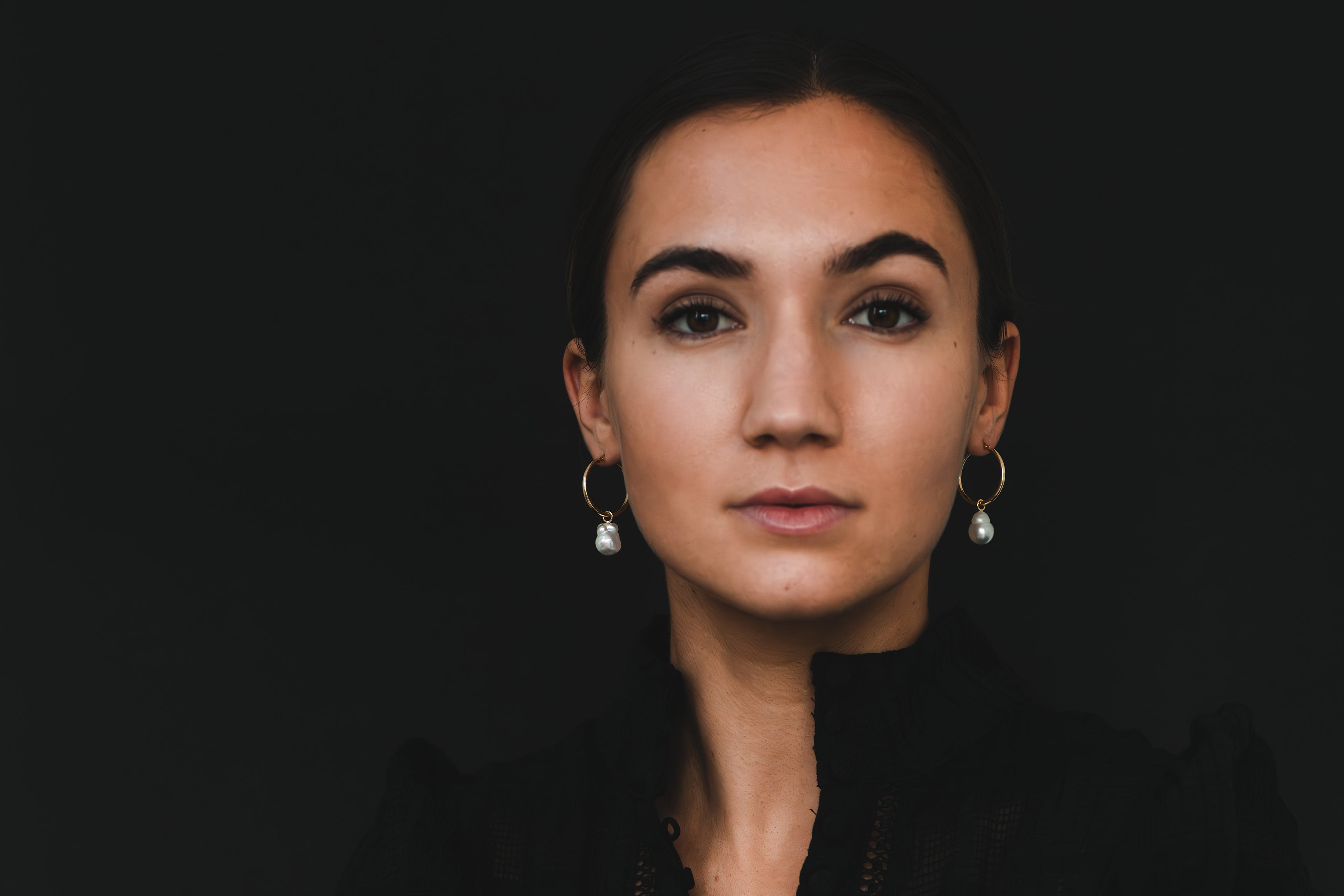 A woman wearing the mini Ayla hoops in yellow gold featuring baroque white South Sea pearls looks directly into the camera.