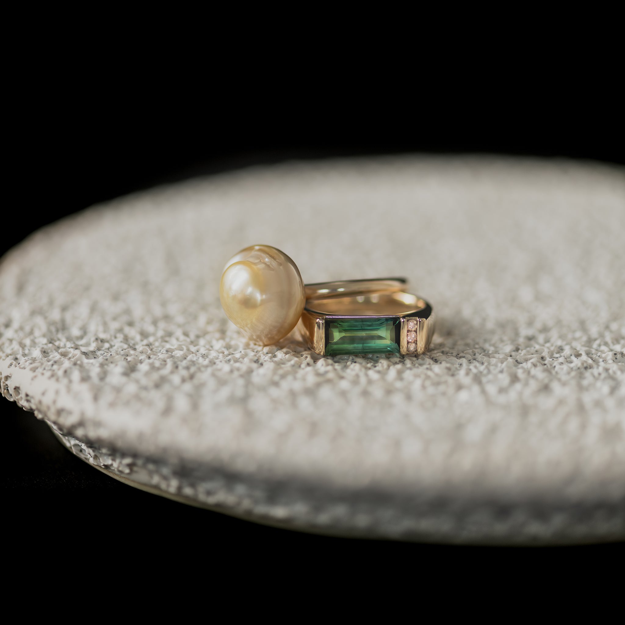 Yellow gold Ayla Pearl Ring with golden baroque South Sea pearl & custom bespoke ring with bi-colour green tourmaline & chocolate Australian champagne diamonds rest on a white textured ceramic dish.