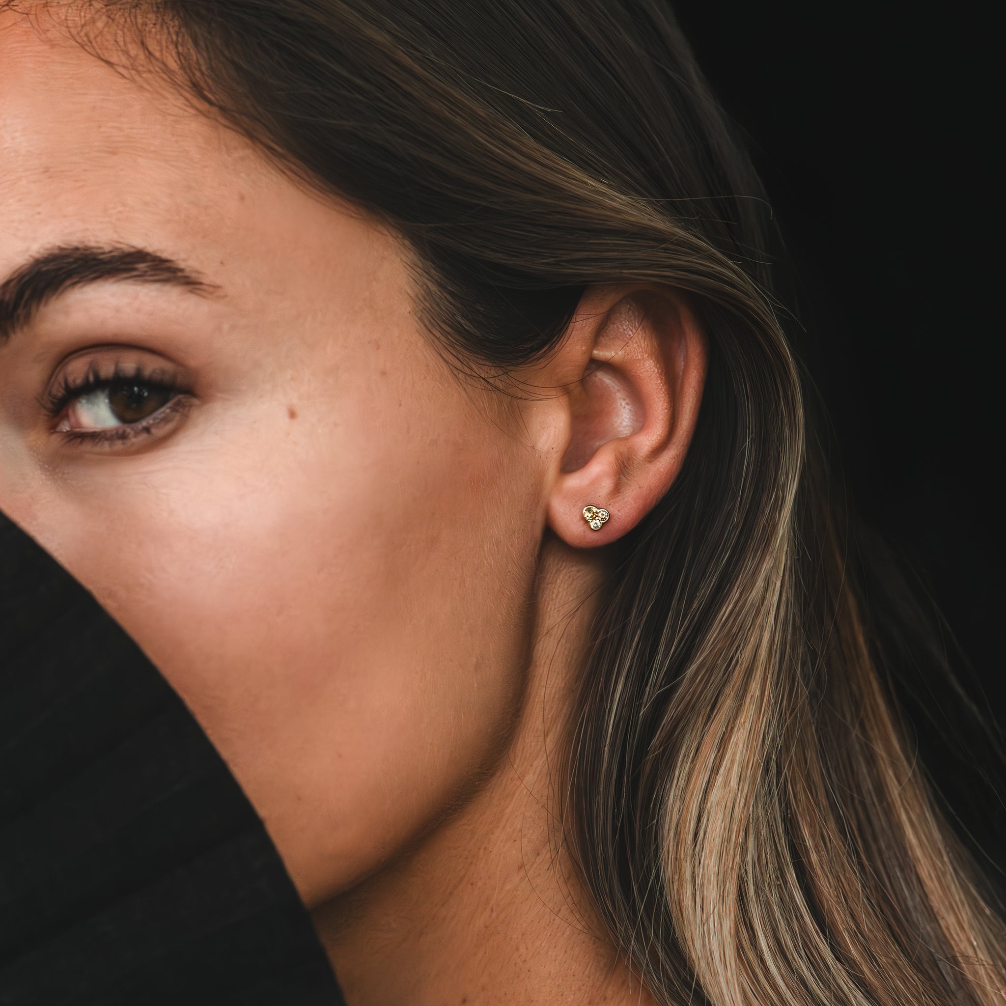 A close up of a yellow gold stud earring featuring a trio of earthy coloured Australian champagne diamonds is worn in the earlobe of a woman who is looking directly into the camera.