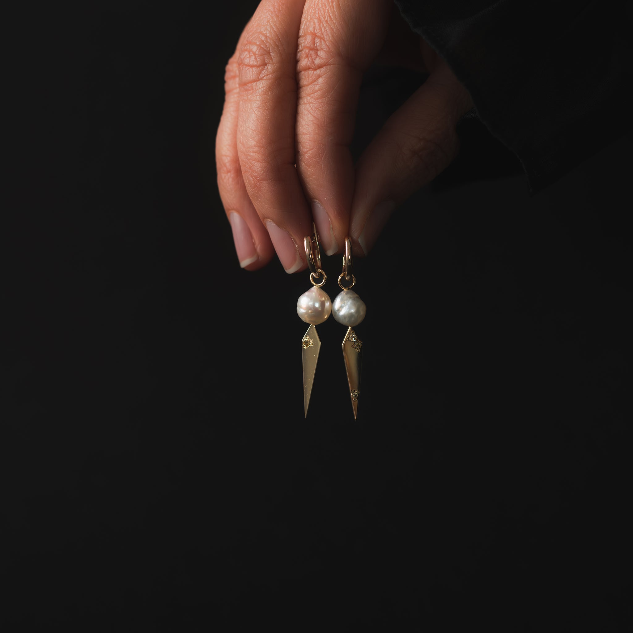 A pair of white baroque South Sea pearl and yellow gold kite shaped drops featuring an earthy mix of Australian champagne diamonds adorning the huggie earrings are being held closely together between a woman&#39;s fingers.