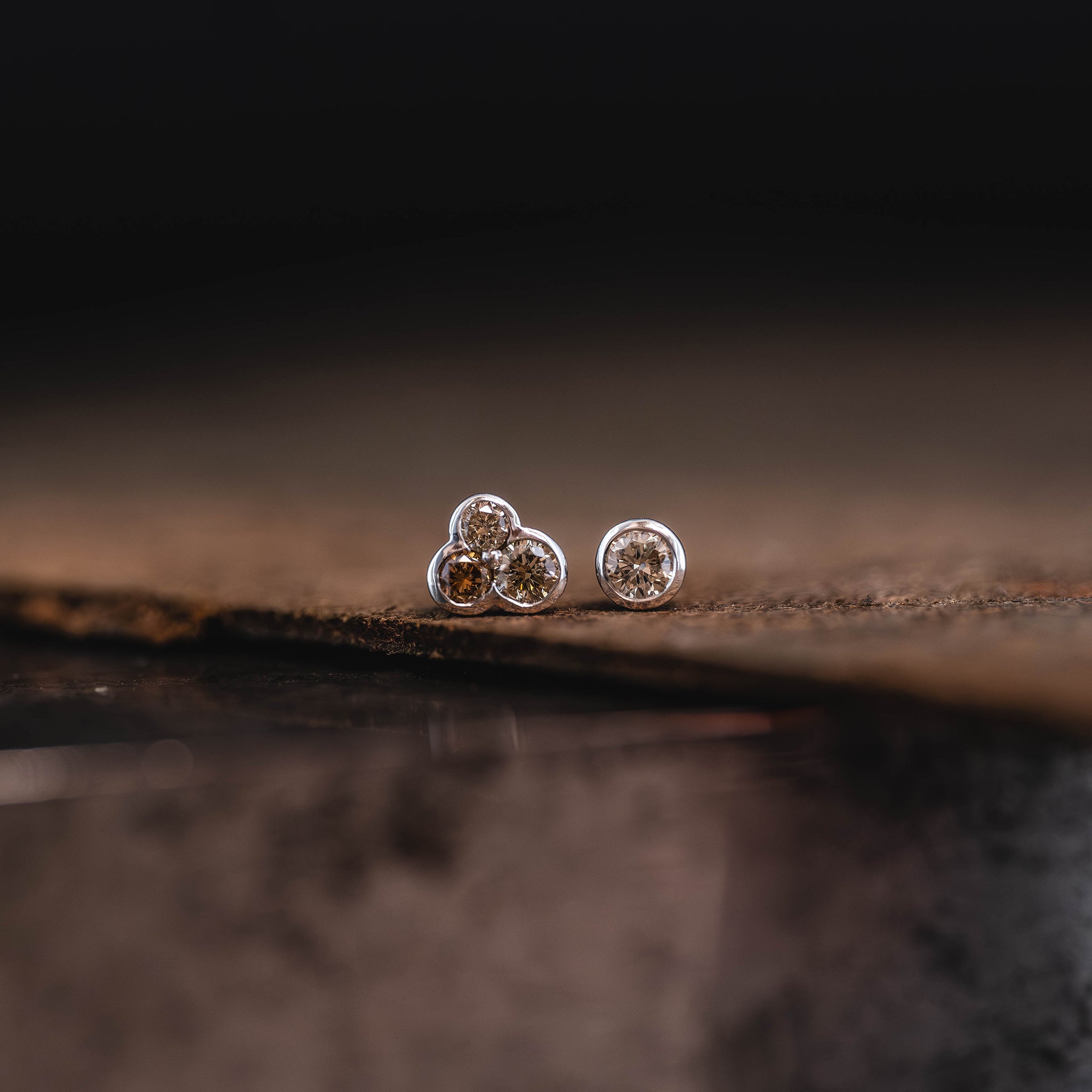 A pair of white gold stud earrings featuring a trio of earthy coloured Australian champagne diamonds in one stud, and a single Australian champagne diamond in the other stud rests on a piece of textured leather.