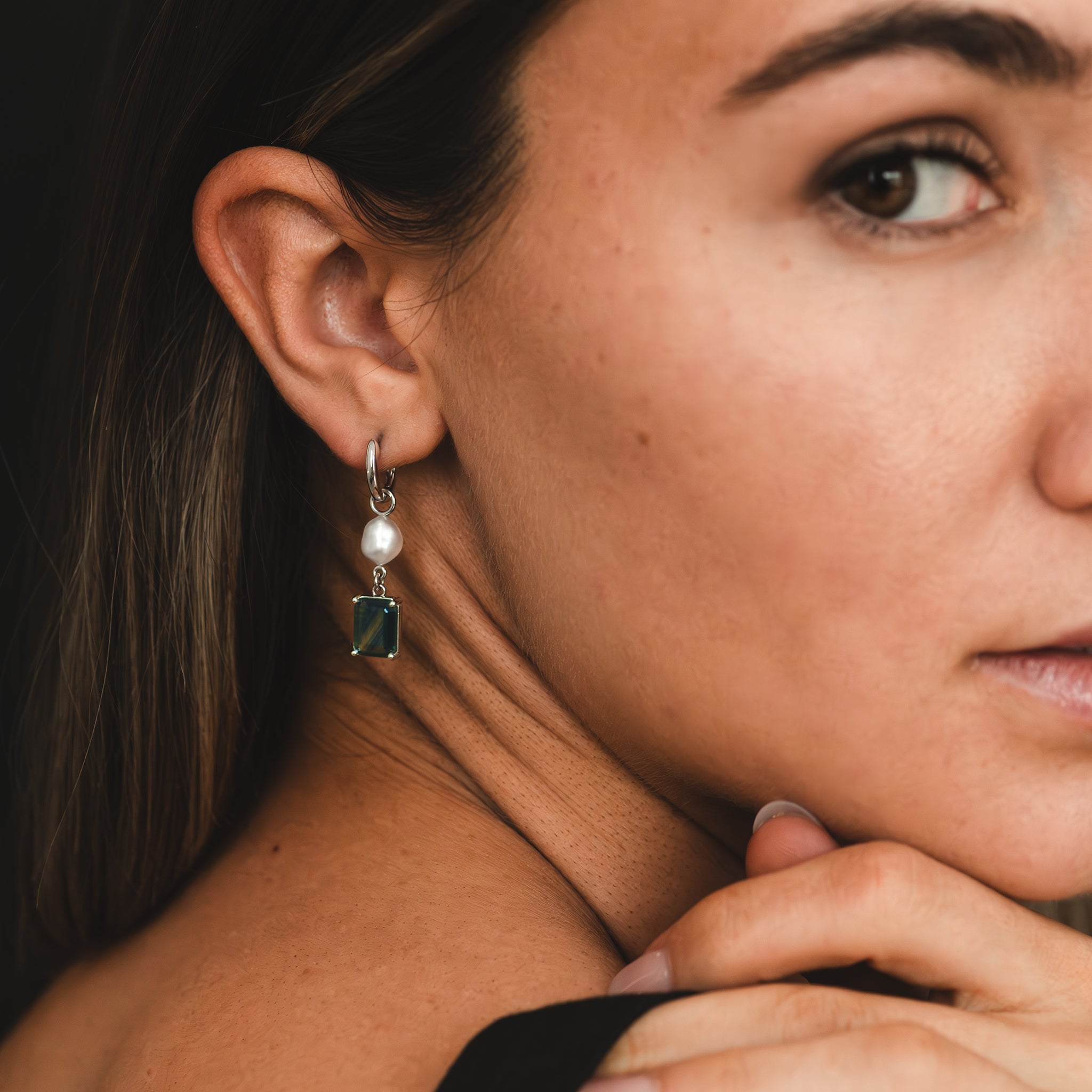 A one-of-a-kind white gold huggie earring featuring a white South Sea pearl and a bi-colour blue Australian sapphire, hand-selected for its captivating, unique colour banding, are adorning a woman's ear lobe.