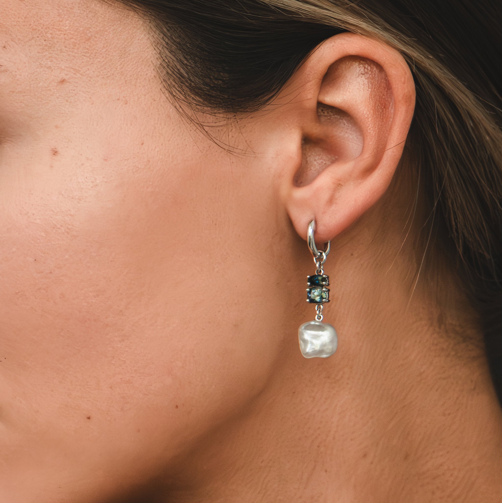 A one-of-a-kind white gold huggie earring featuring a white Keshi pearl and a medley of bi-colour blue Australian sapphires, all hand-selected for their captivating, unique colour banding, are adorning a woman's ear lobe.