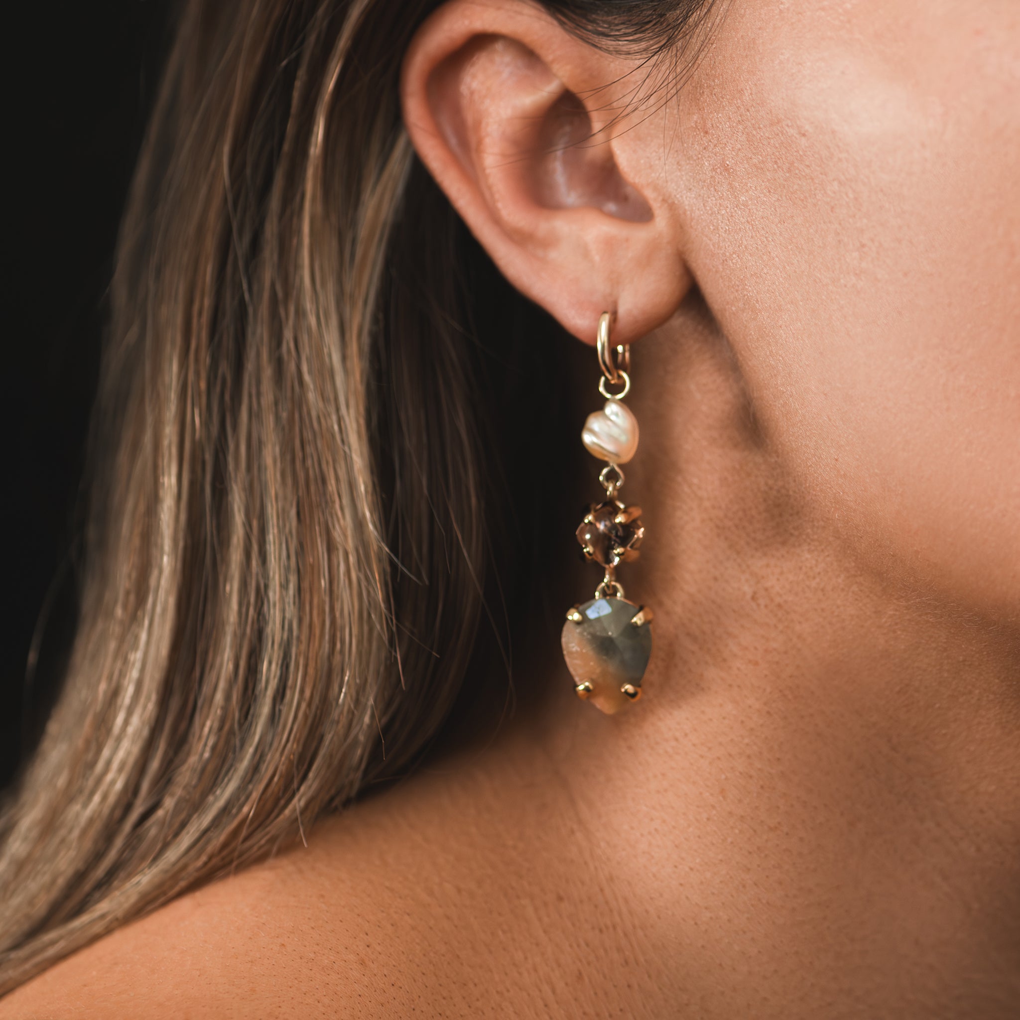 One-of-a-kind rose-cut peach and khaki sapphires, a soft cream Keshi pearl and gold-toned tourmaline cabochon drop adorning yellow gold huggie earrings are adorning a woman's ear lobe.