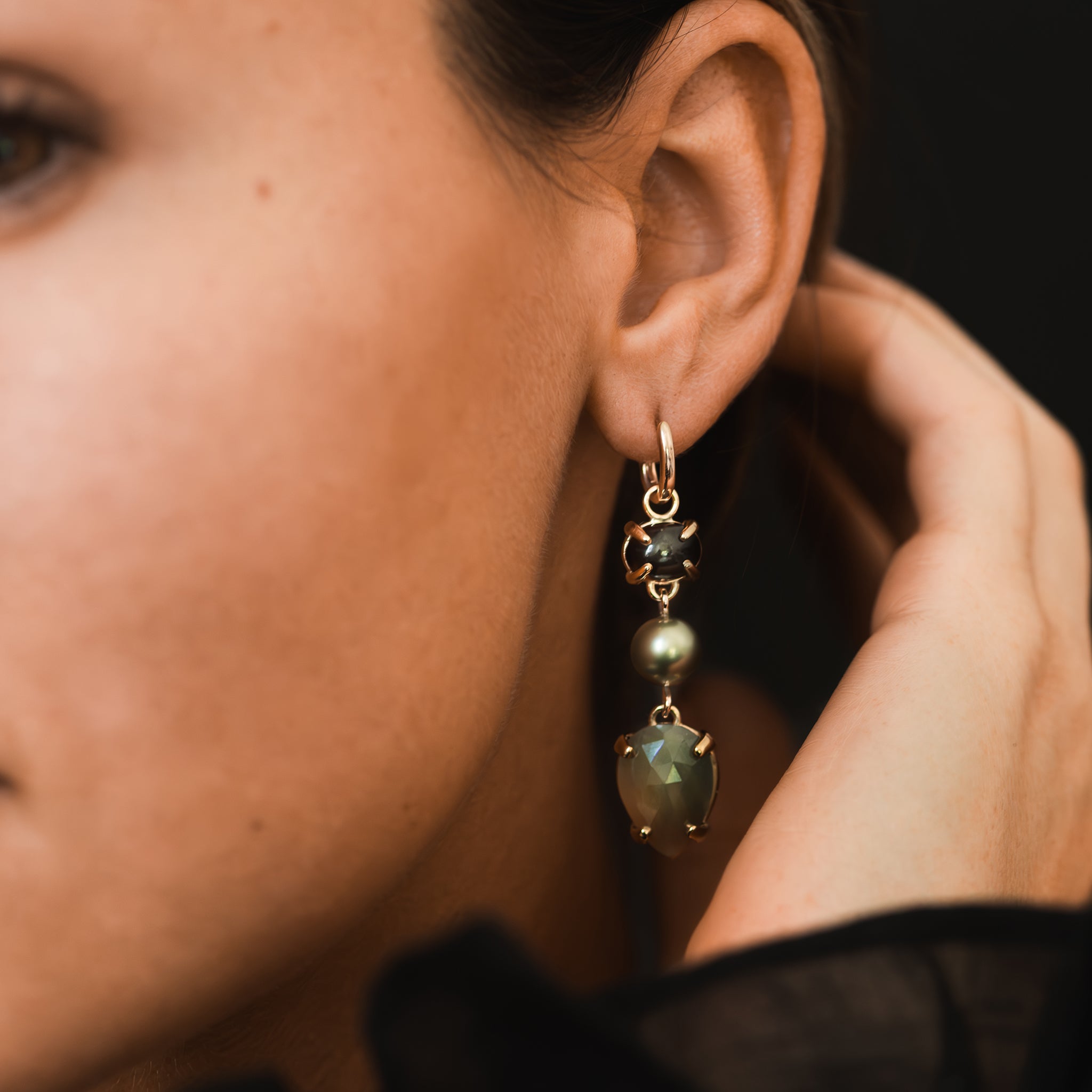 One-of-a-kind rose-cut peach and khaki sapphires, a pistachio-hued Tahitian pearl, and an olive-toned tourmaline cabochon drop yellow gold huggie earrings are adorning a woman&#39;s ear lobe.