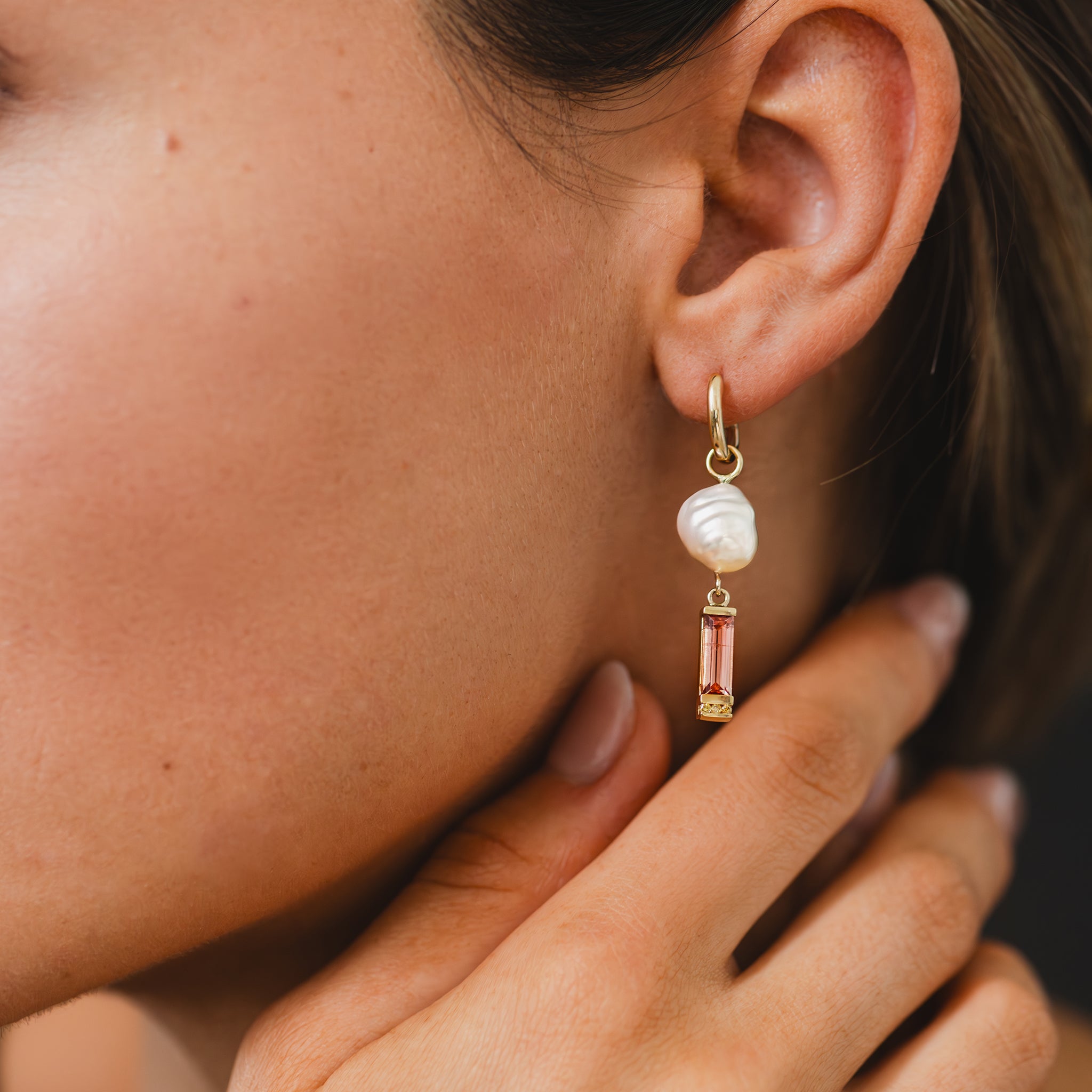 A close up of the one-of-a-kind soft pink tourmaline, Australian champagne diamond, and South Sea Keshi pearl drop, yellow gold, huggie earrings adorning a woman&#39;s earlobe.